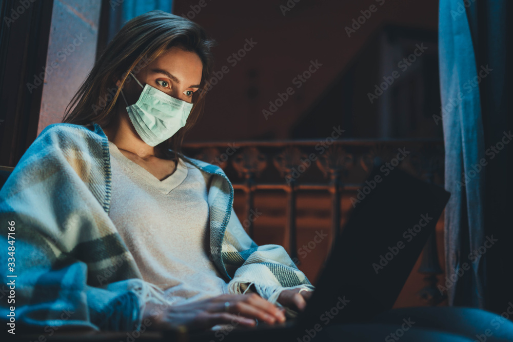 Young woman wearing face mask during coronavirus and flu outbreak working at home at night using laptop computer, sick girl in medical mask using portable computer browsing internet or social networks