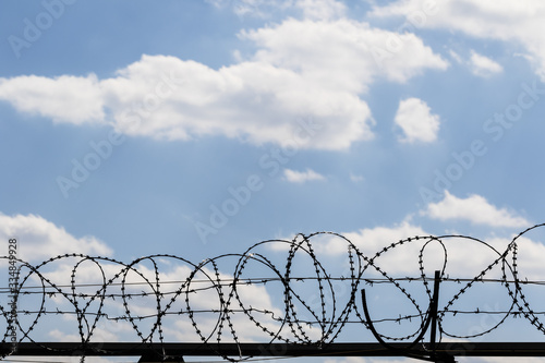 Black coiled barbed wire fence and blue sky with sunlit clouds