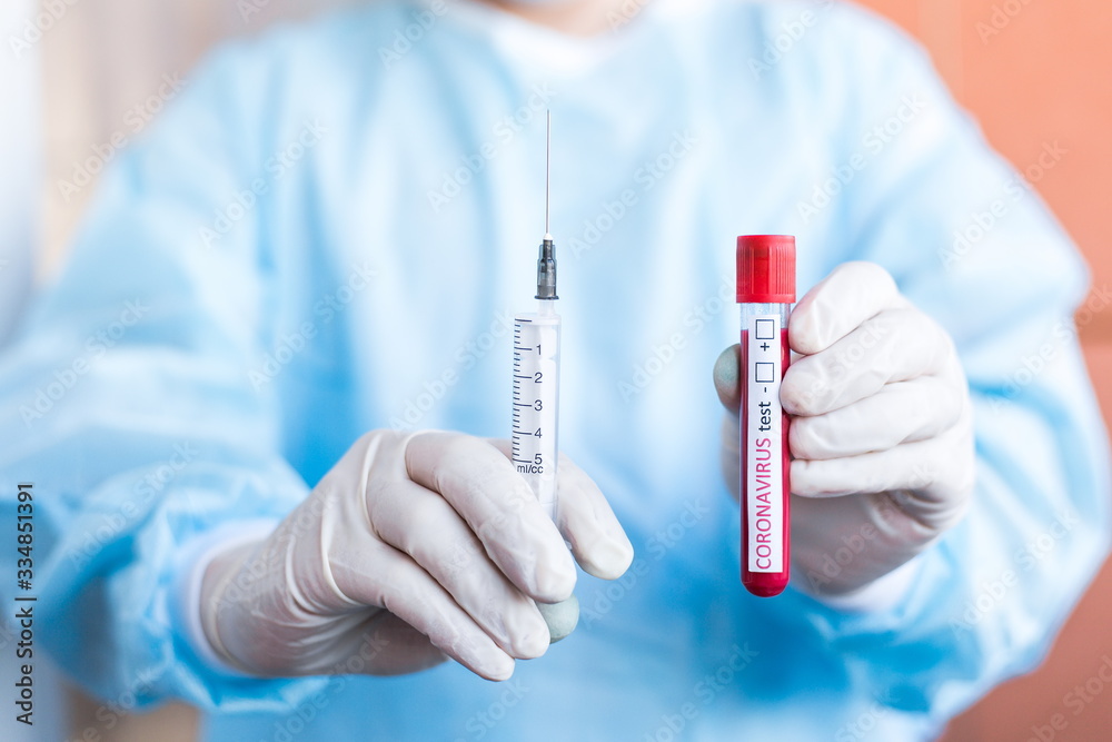 Doctor standing in a protective suit. Holding a medical tube containing a blood sample for testing COVID-19 with a syringe Doctor standing in a protective suit. Holding a medical tube containing a blood sample for testing COVID-19 with a syringe