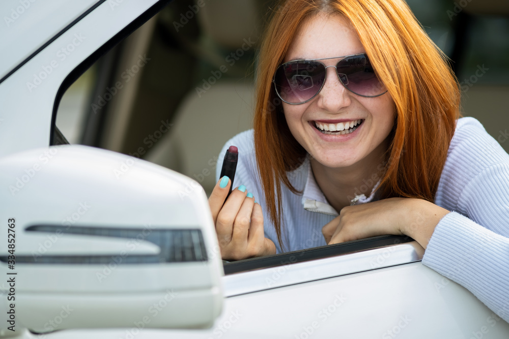 Closeup of a young redhead woman driver correcting her makeup with dark red lipstick looking in car rearview mirror behind steering wheel of a vehicle.