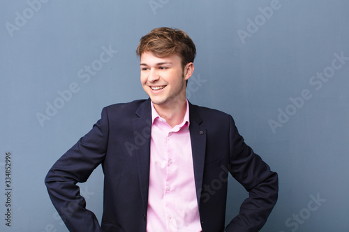 young blonde man looking happy, cheerful and confident, smiling proudly and looking to side with both hands on hips isolated against flat wall photo