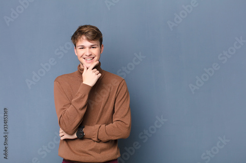 young blonde man smiling, enjoying life, feeling happy, friendly, satisfied and carefree with hand on chin isolated against flat wall photo