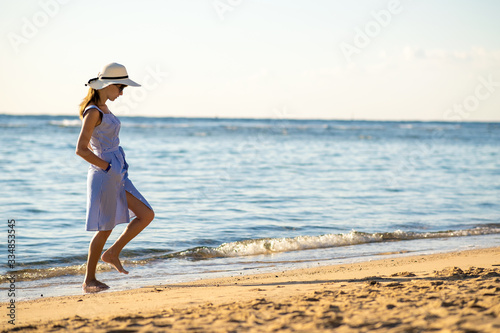 Young woman in straw hat and a dress walking alone on empty sand beach at sea shore. Lonely tourist girl looking at horizon over calm ocean surface on vacation trip.