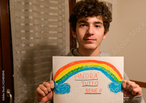 Turin, Piedmont, Italy. March 2020. Cornonavirus quarantine. Portrait of a brown-haired caucasian curly-haired boy holding up a drawing with the rainbow and the message will be all right! photo