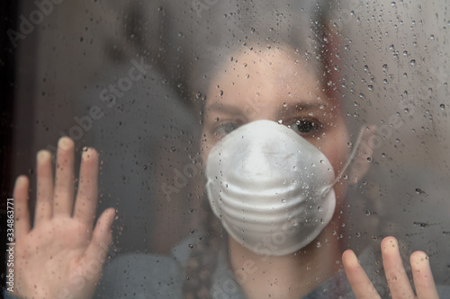 COVID-19 Pandemic Coronavirus, Young girl in home isolation, during quarantine is wearing protective face mask for stave off preading of disease virus, looking out of the window photo