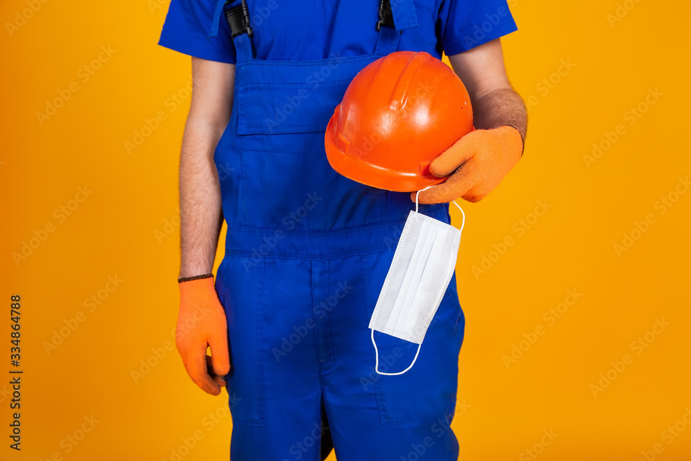 Worker in construction overalls and gloves on a yellow background, holds a protective helmet and a medical mask in his hand. The concept of the economic crisis, unemployment production, coronavirus Worker in construction overalls and gloves on a yellow background, holds a protective helmet and a medical mask in his hand. The concept of the economic crisis, unemployment production, coronavirus