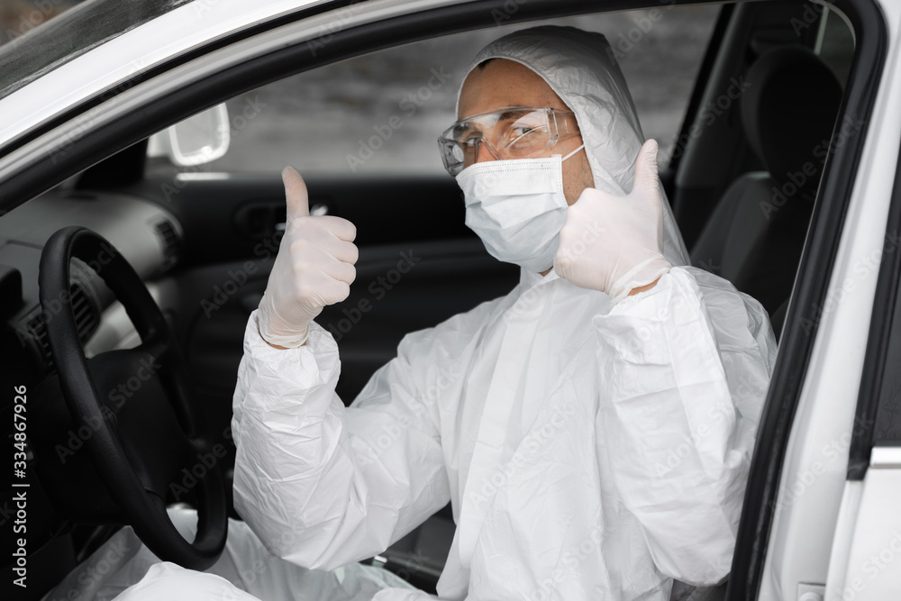 Man in protective suit, medical mask and rubber gloves for protect from bacteria and virus is sitting in a car and showing a thumb up. Quarantine, world pandemic, covid 19, coronavirus, infection. Man in protective suit, medical mask and rubber gloves for protect from bacteria and virus is sitting in a car and showing a thumb up. Quarantine, world pandemic, covid 19, coronavirus, infection.