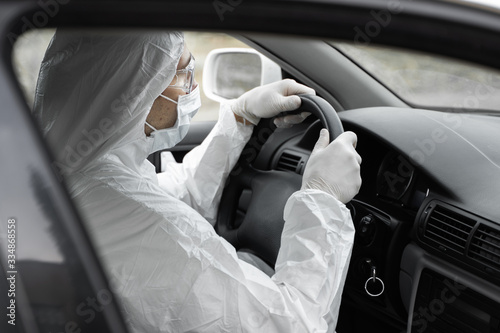 Man in protective suit, medical mask and rubber gloves for protect from bacteria and virus is driving a car. Protective mask while quarantine, world pandemic, covid 19, coronavirus, infection. © Volodymyr