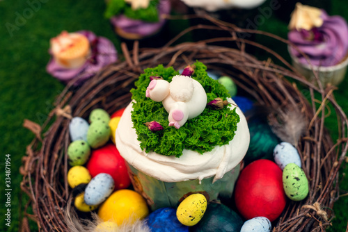 Orthodox Easter cake in a wooden nest with painted chicken and quail eggs on green grass. Easter cake with a figurine of a ponytail of a rabbit on the background of sweets, cupcakes