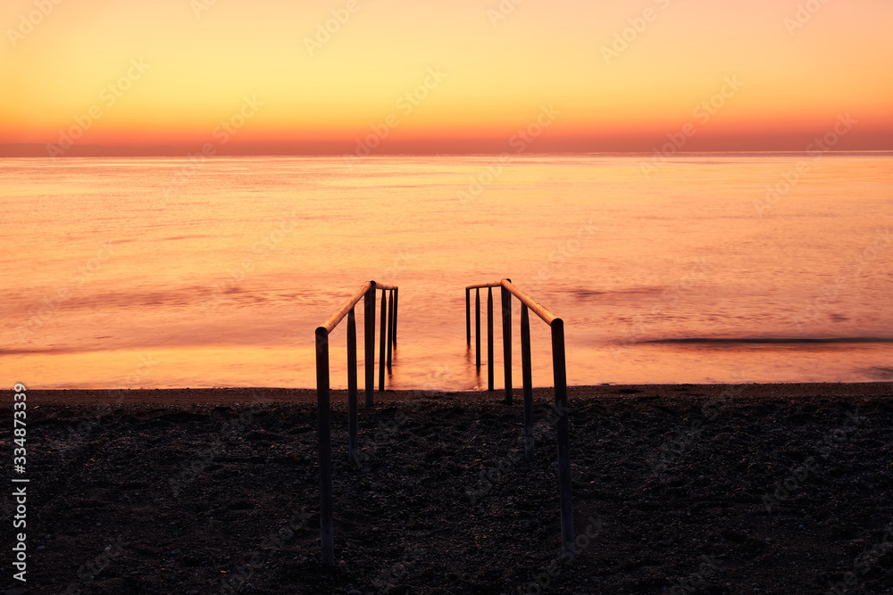 Beautiful warm sea and stairs on the beach for entering the water against the background of orange dawn.