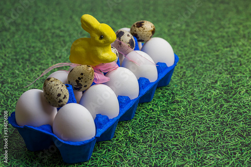 white chicken and quail eggs in a blue uniform, a tray and a yellow Easter bunny with a pink bow on a background of green grass