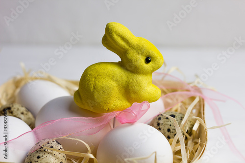 white large chicken and quail eggs on a straw in a basket with a yellow easter bunny and a pink leto straw on a white light wooden background