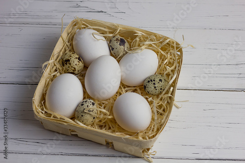 white large chicken and quail eggs in a basket with straw on a white light wooden background
