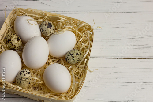 white large chicken and quail eggs in a basket with straw on a white light wooden background