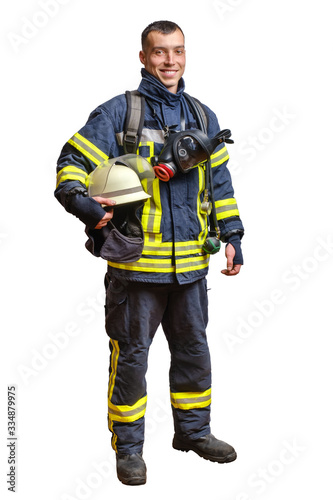 young smiling fireman in a fireproof uniform stands and looks at the camera with a helmet in his hands.