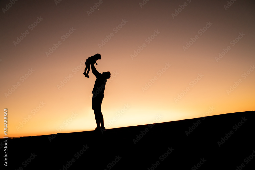 Father's day. Silhouette of a happy joyful father having fun throws up in the air little cute child baby girl on nature, sunset horizon background.