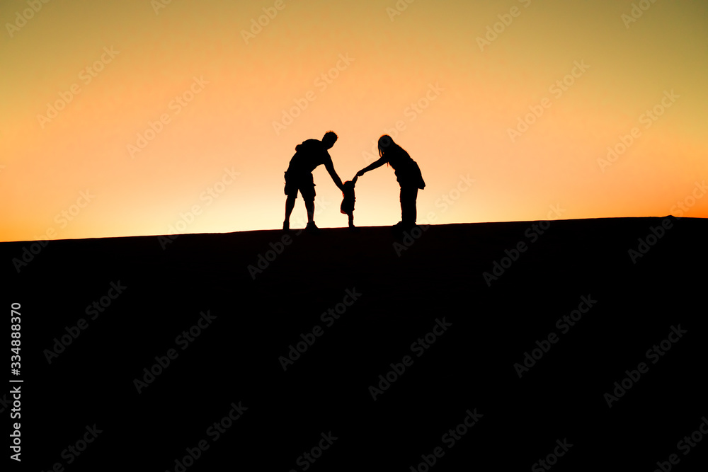 Silhouette of a happy joyful family having fun holding hands with their little cute child baby girl on desert, sunset horizon background.