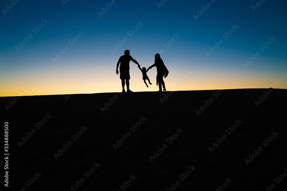 Silhouette of a happy joyful family having fun holding hands with their little cute child baby girl on desert, sunset horizon background.