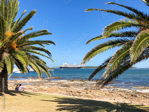 Young couple sitting in shadow of palm tree watch luxury Azamara Club Cruises cruiseship or cruise ship liner Azamara Pursuit anchor in bay in front of Punta del Este in Uruguay in summer photo