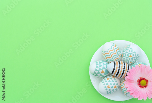 Decorated Easter eggs lie on the white plate with pink flower on green background. Happy Easter holiday and spring concept. Greeting  invitation card. Flat lay style with copy space.