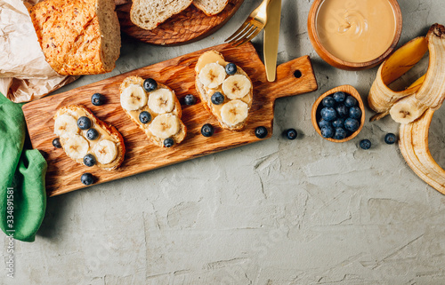 Buckwheat healthy bread with peanut butter, banana and blueberry on wooden board over concrete background. Top view. Flat lay. Summer breakfast.