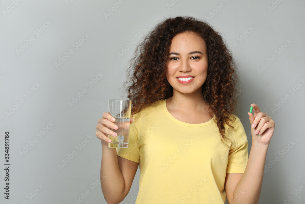 African-American woman with glass of water and vitamin capsule on light grey background African-American woman with glass of water and vitamin capsule on light grey background