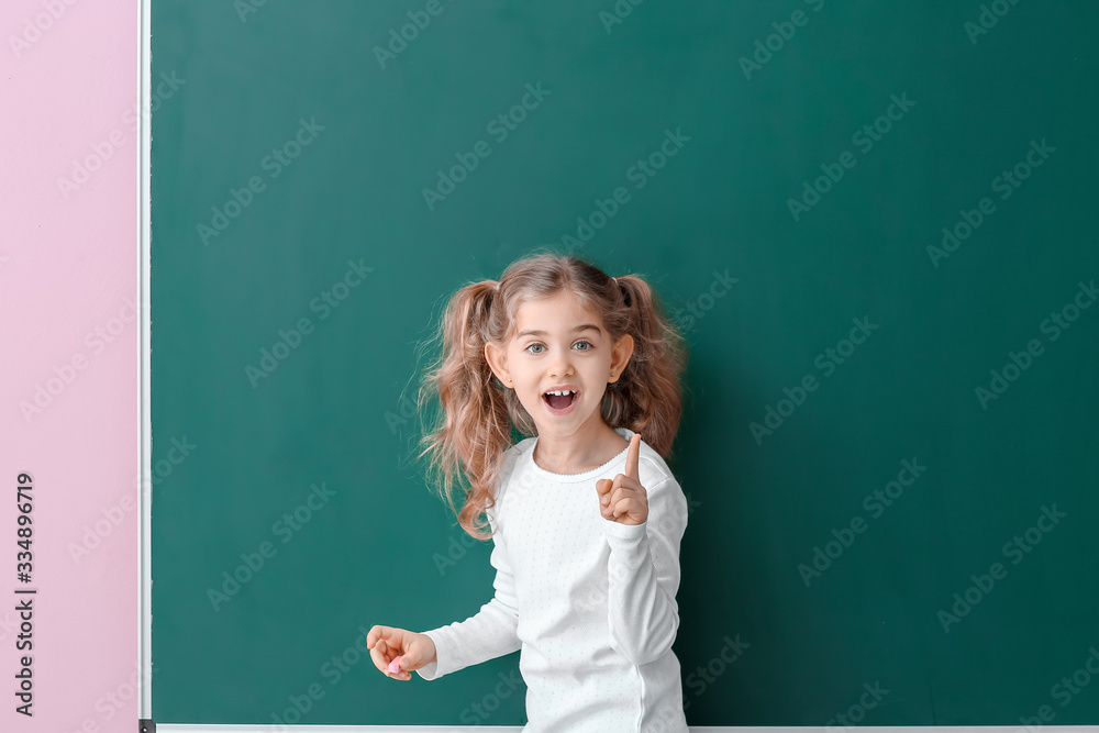 Cute little schoolgirl with raised index finger near blackboard in classroom