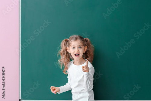 Cute little schoolgirl with raised index finger near blackboard in classroom