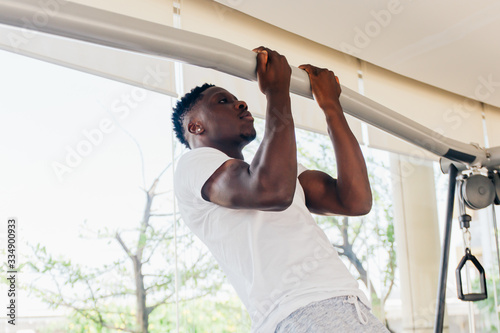 Strong African American sportsman in white T-shirt doing pull ups during fitness workout in modern gym.