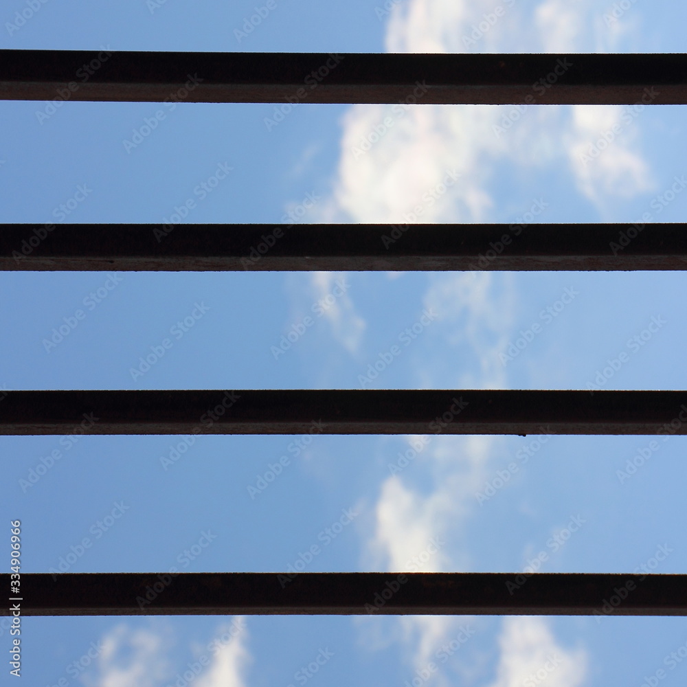 Fototapeta premium Blue sky with clouds, view through grating horizontal iron bars - self-isolation concept blank for text, quarantine, remote work from home