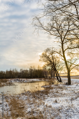 The orange sun behind the trees, the fields with a pond and tall grass covered in snow. Beautiful sunset in early spring sun with blue sky and clouds.