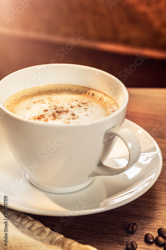 Eine Kaffeetasse mit ger  steten Kaffeebohnen auf einem h  lzernenen Tisch. Sonneneinstrahlung  Reflexionen. Ein Cappuccino mit Milchschaum und gemahlenem Kaffee. Serviert in einem Caf  . Milchkaffee