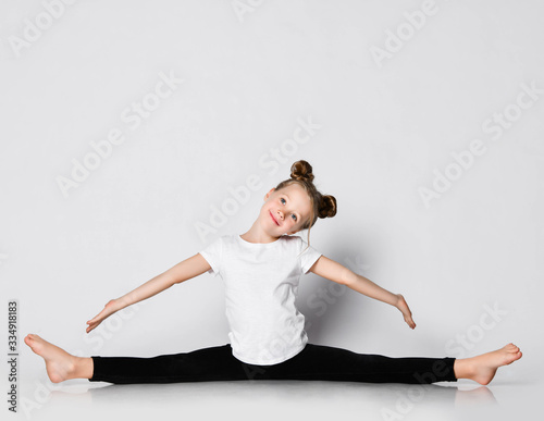 Smiling kid girl does gymnastic exercises at home splits with her arms outstretched and head tilted aside