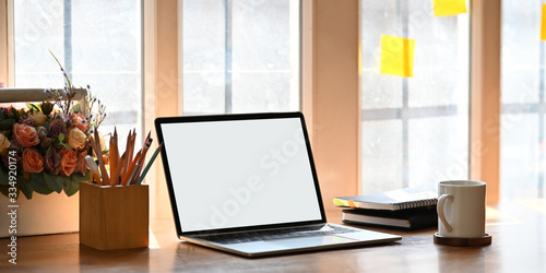 Photo of computer laptop with white blank screen putting wooden working desk with pencil holder, stack of books, coffee cup and basket of flowers over modern and comfortable workplace as background. © Prathankarnpap
