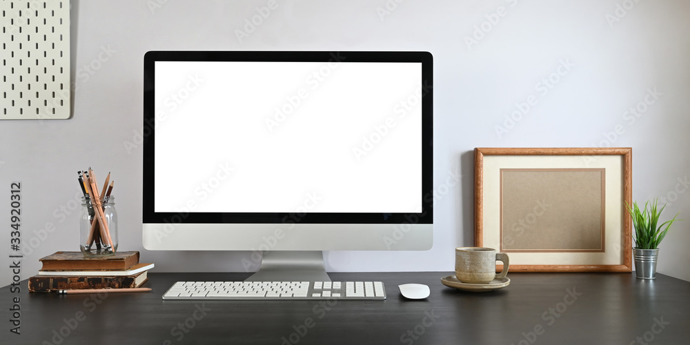 Photo of Computer monitor with white blank screen putting on working desk with wireless mouse, keyboard, pencil holder, stack of books, coffee cup and potted plant over white wall as background. Photo of Computer monitor with white blank screen putting on working desk with wireless mouse, keyboard, pencil holder, stack of books, coffee cup and potted plant over white wall as background.