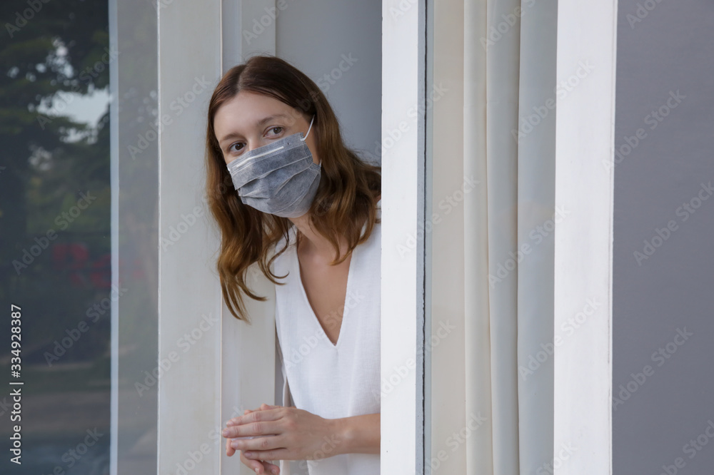 Young woman with surgical disposable mask looking outside through window. Bored and completely alone at home. Coronavirus outbreak and self-quarantining concept.