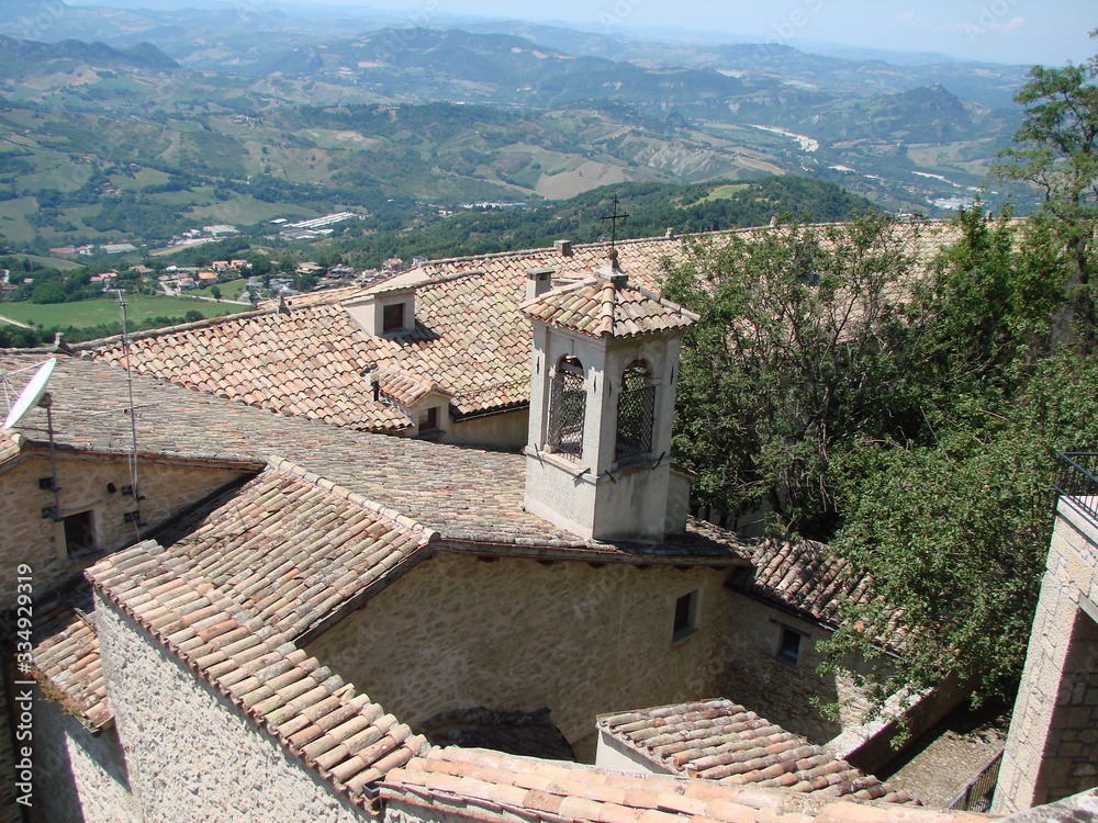 View from above of the tiled roofs of ancient houses of a small state under the rays of the summer sun surrounded by garden trees.