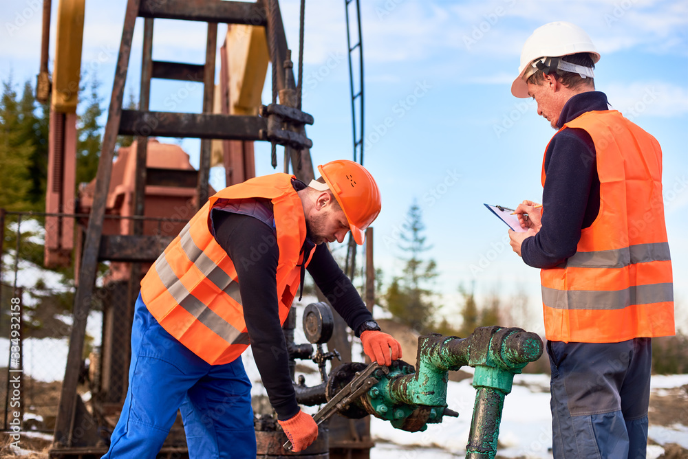 Two oil workers wearing overalls, orange vests, and helmets, working on oil field next to oil pump jack, one is wrenching the pipe, the other making notes. Concept of petroleum industry, collaboration Two oil workers wearing overalls, orange vests, and helmets, working on oil field next to oil pump jack, one is wrenching the pipe, the other making notes. Concept of petroleum industry, collaboration