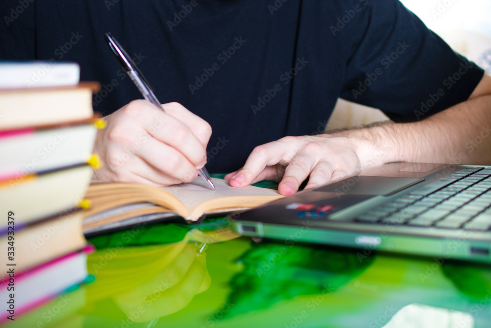 Learning at home, hands writing with a pen in a notebook, a person sitting at a table and studying or working on a laptop against the background of books Learning at home, hands writing with a pen in a notebook, a person sitting at a table and studying or working on a laptop against the background of books