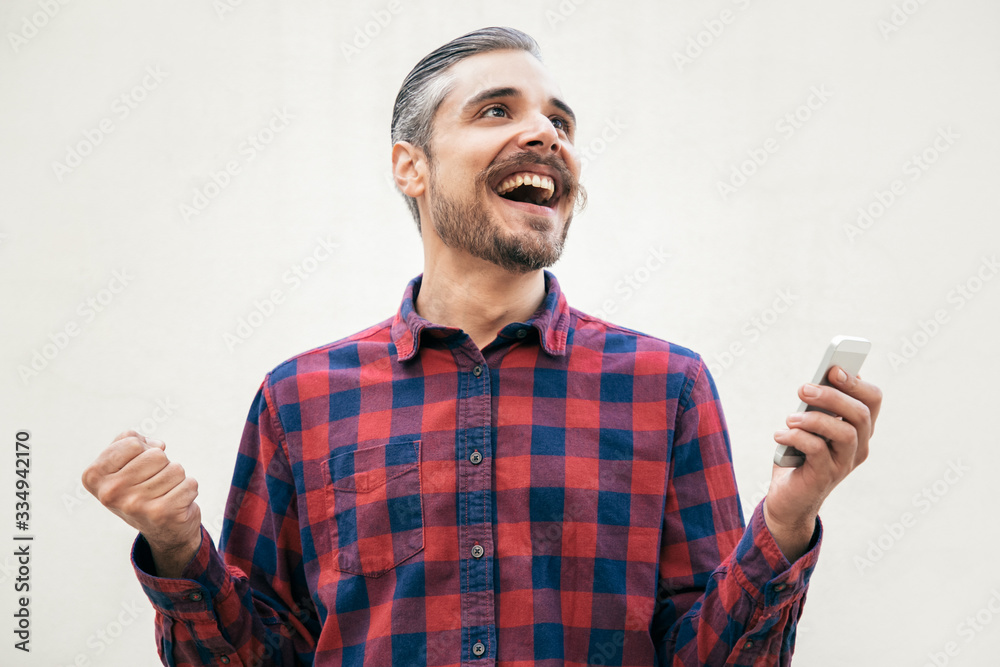 Overjoyed excited man with cellphone making winner gesture. Grey haired young man in checkered casual shirt posing isolated over white background. Celebrating win or success concept Overjoyed excited man with cellphone making winner gesture. Grey haired young man in checkered casual shirt posing isolated over white background. Celebrating win or success concept