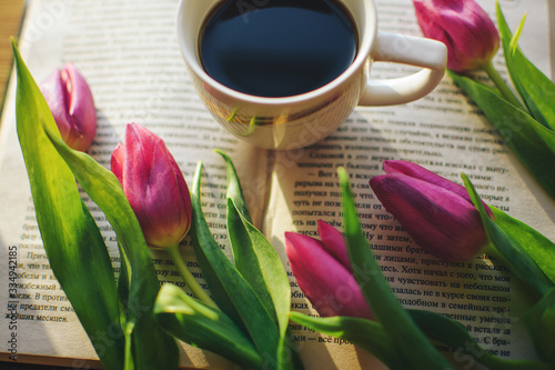 Strong tasty aromatic coffee on the window with an interesting book and pink tulips. The morning sun shines on a book and aromatic coffee, and beautiful fresh pink tulips lie nearby
