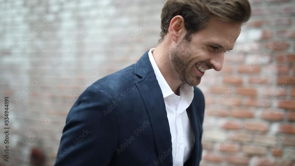 elegant businessman wearing a navy suit is looking around and walking forward then he is turning around, moving backward and fixing his jacket near a brick wall