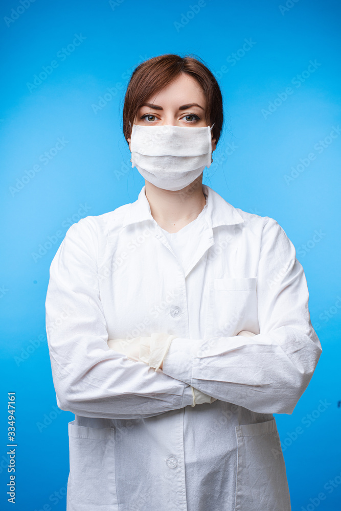 Portrait of professional female doctor in white uniform and protective mask posing isolated. Practitioner woman medical personnel in safety suit stand with crossed hands on blue studio background Portrait of professional female doctor in white uniform and protective mask posing isolated. Practitioner woman medical personnel in safety suit stand with crossed hands on blue studio background