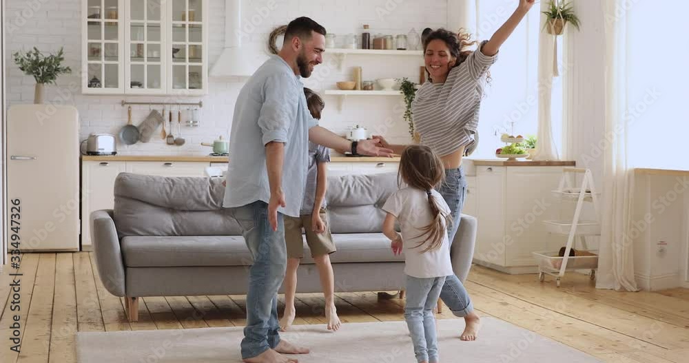Happy active family of four young parents and cute small children jumping together in kitchen interior, carefree funny little kids with father mother having fun dancing laughing bonding at home