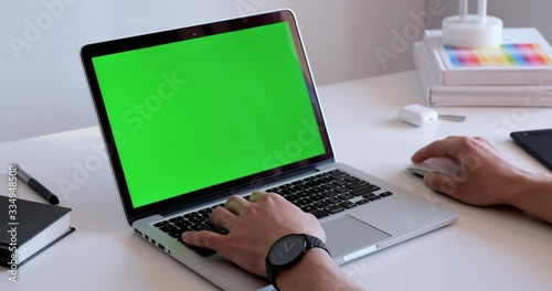 Man working in front of a laptop in a white modern office with a green screen chroma key display. Hands typing on a keyboard, using a wireless mouse on a clean desk with office tech gadgets and books