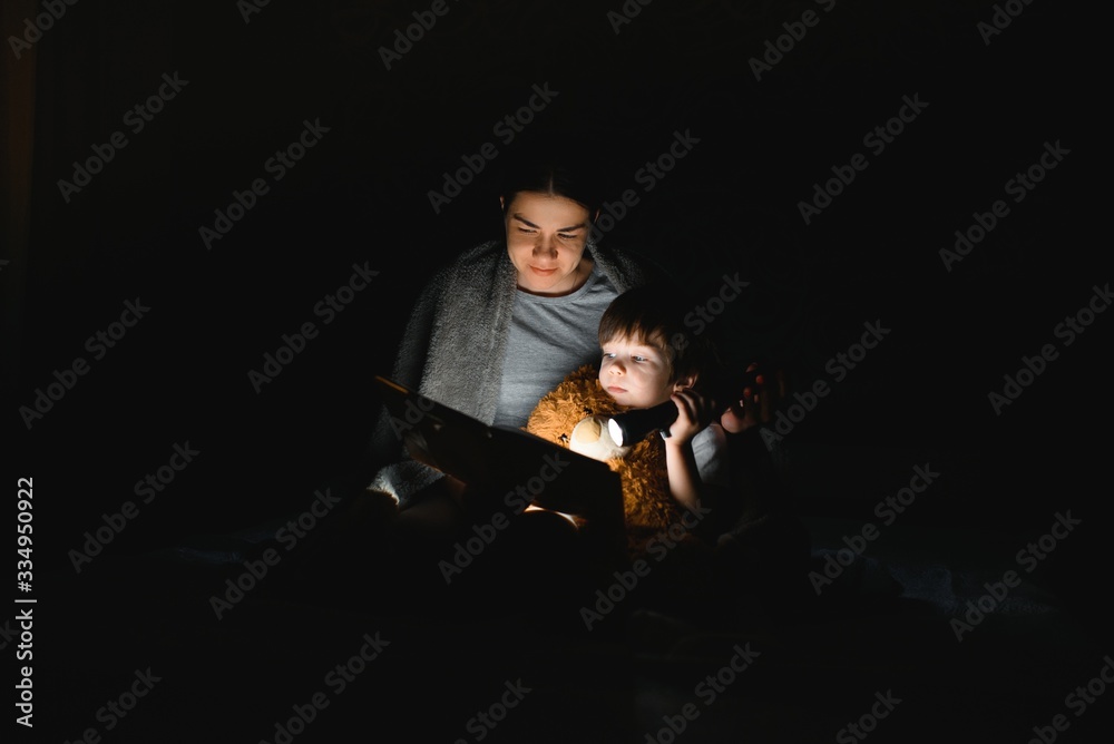 Family Mom and little son reading a book with a flashlight under the blanket.