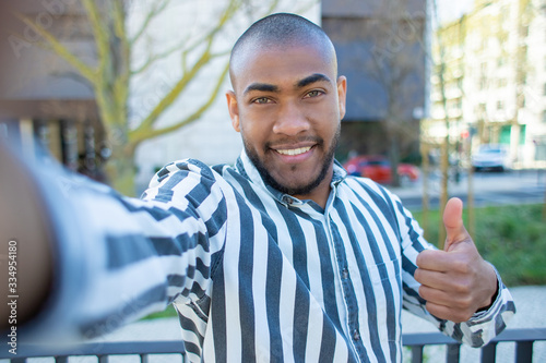 Handsome smiling African American man showing thumb up to camera. Cheerful young guy holding digital device in outstretched arm while posing for selfie. View from camera. Self portrait concept