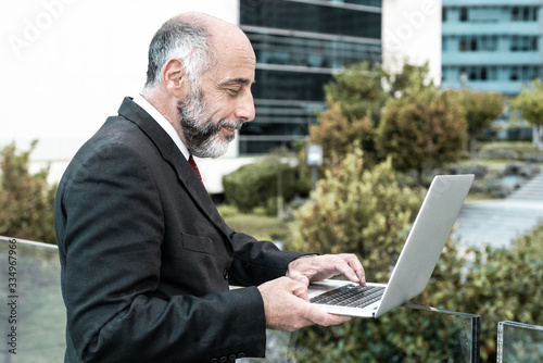 Side of positive mature business leader working with laptop near office building. Elderly man in formal suit and tie walking outside in city. Digital communication concept © Mangostar