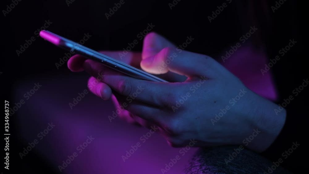 Young woman texting chatting on smart phone at night; girls finger tapping keypad, writing message on mobile devices touchscreen, blue low light; close up top side view