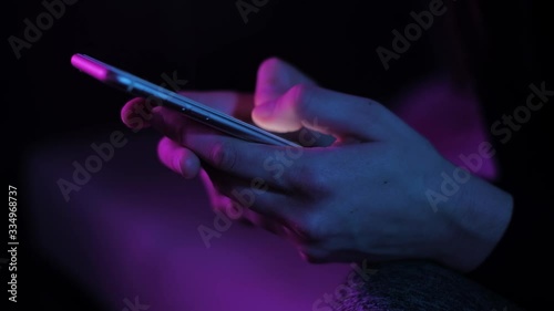 Young woman texting chatting on smart phone at night; girls finger tapping keypad, writing message on mobile devices touchscreen, blue low light; close up top side view